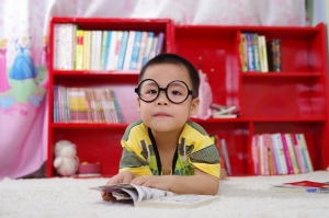 Kid with big round blag glasses looking at the camera and holding a book